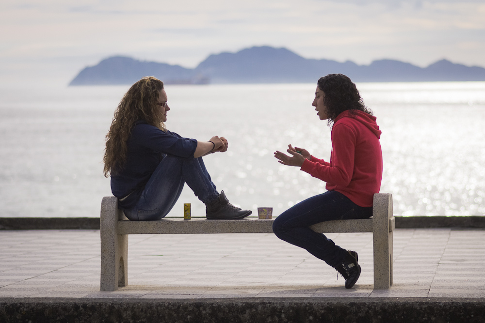 Dos chicas hablan en un banco ante un paisaje de mar y montañas